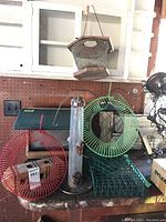 Full view of various bird feeders on table showing red and green circular peanut feeders, clear tube feeder, green rectangular feeders, and the Wild Bird Unlimited resin feeder hanging above