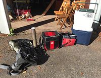 Image shows two Coleman coolers stacked, one white and green with wheels and one blue and white, along with two red and black soft cooler bags, a gray and black hiking backpack, and a portable chess set on the ground nearby.