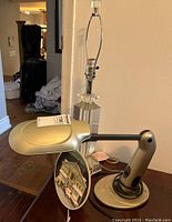 Desk lamp with metallic arm and lighted head angled forward, cut glass lamp base without shade behind, lighted resin Norman Rockwell plate leaning against base.
