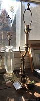Two table lamps standing side by side on a windowsill, shadows from window showing. Both lamps have glass bases and brass fittings, without shades. One extra harp is visible in the lot.