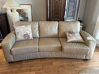 Front view of three-seat beige leather sofa with two floral accent cushions on hardwood floor.