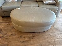Overall front view of tan leather ottoman on hardwood floor with sofa in background.
