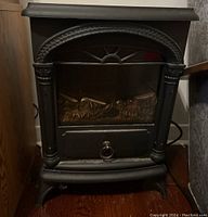 Front view of black metal portable electric fireplace heater showing decorative arch and columns, window with ring handle.