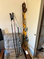 Photo showing full view of Salomon skis leaning against wall with bindings, Ultra poles standing beside skis, Easton bat resting on chair, and Oakley goggles hanging on skis.