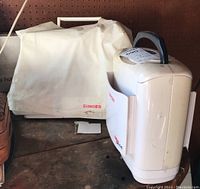 Singer sewing machine covered with a white dust cover labeled Singer next to a white hard accessory case marked Singer.