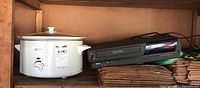 Photo showing white Betty Crocker slow cooker and black RCA VCR side by side on a wooden shelf.