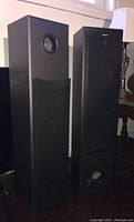 Two black Sony SS-MF415 floor tower speakers standing side by side on floor under window. One speaker has grille attached, the other without grille showing drivers.