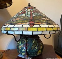 Close-up view of the stained glass lamp shade and base showing dragonfly design and colorful glass pieces with electrical cord and plug visible.