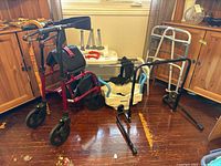 General overview of the lot showing rollator, carved cane, transfer bench, dual assist handle, and walkers arranged in a room corner.