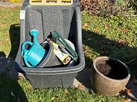 Photo of black plastic wheelbarrow containing turquoise watering can, black planter, and gardening tools. Brown planter beside it on ground.