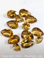 Photo showing 12 faceted genuine citrine gemstones in yellow-golden hues arranged on a white surface.