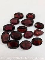 Close-up image showing a collection of loose garnet gemstones with various polished cuts including oval, round, and pear shapes. Deep red color with good clarity and faceted surfaces.