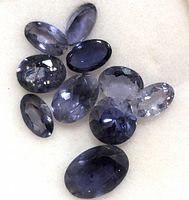 Loose, faceted iolite gemstones with oval cuts showing violet-blue color scattered on a white surface.