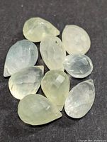 Photo showing 10 pear-shaped faceted phrenite gemstones in pale yellow and light green hues, arranged on a dark background.