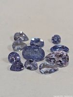 Photo showing multiple loose genuine tanzanite gemstones in various cuts and sizes with blue to violet coloration, laid out on a neutral surface.