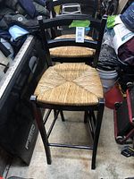 Two black wooden bar stools with woven seats placed side by side in a cluttered storage area. Both stools have matching design and show minor signs of use.