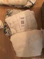 Photo of folded bedding pieces including a white and blue grid pattern sheet, a floral patterned piece and plain white bedding on floor.