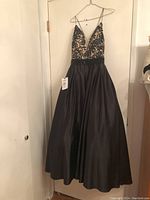 Front view of the black formal gown with lace and satin fabric, hanging on a hanger against a white door.