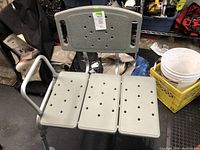 Photo showing a light gray plastic three-panel shower transfer bench with multiple drainage holes and a single armrest on the left side. The bench has adjustable metal legs and is shown in a cluttered background.