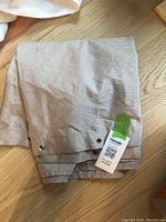 Photo of folded beige shower curtain showing perforated sunburst pattern and metal grommets with a MaxSold tag attached.