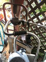 Photo showing rusted metal weed wacker frame with black rubber handles and grips leaning on lattice fence.