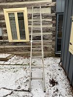 Full view of the 24 feet extendable metal ladder leaning against a log cabin wall outdoors, showing the ladder at full extension with a yellow-framed window in the background.