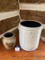 Both crocks placed on wooden surface against rustic wall; large white crock marked '8' is prominently visible, medium crock beside it.