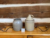 Both ceramic crocks displayed side by side on a wooden shelf with metal brackets, showing size and condition.