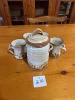 Photo showing four ceramic coffee mugs with matching ceramic jar with lid on a wooden table.
