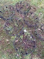 Six black wire-frame hanging plant baskets with chains, arranged on grass. Baskets are circular with spoke-like supports, black painted metal, some chain rust visible.