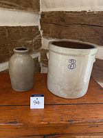 Photo showing both crocks on a wooden surface in front of a rustic wooden wall, large crock with number 3 visible