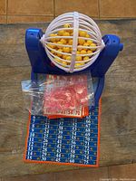 Photo of Bingo game with blue mechanical cage, yellow balls inside, orange numbered cards, and red tokens in bag.