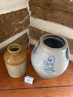 Wide view of two crocks on a wooden floor against wood paneled wall, showing overall size and shape of each.