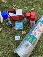 Overview of all items on grass showing the red rotary tumbler, three red/yellow cans, one blue can, suet feeder, and rolled aluminum ducting.
