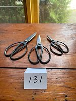 All three black metal scissors of different sizes arranged on wooden surface with lot number card.