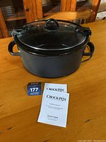 Large black Crock-Pot slow cooker on wooden table with glass lid and printed manuals beside it.