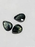 Three pear shaped sapphires showing dark green/blue hues, polished and faceted, lying on a white background, photo taken to display color and shape.