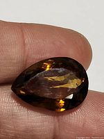 Pear shaped cut dark orange-brown citrine gemstone weighing 17.16ct, held between fingers to show size and color.