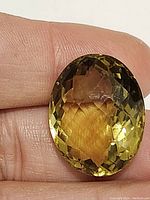 Oval checkerboard cut citrine gemstone held between fingers, showing size and color variation from champagne yellow to light brown.