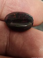 Oval black opal held between fingers showing the play of color with red and green flashes under light.