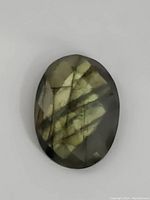 Oval faceted labradorite gemstone with visible labradorescence in green and gold tones on a gray base.