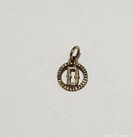 Single 10K gold pendant showing front of circular charm with letter 'B' inside decorative border.