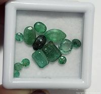 Top view of 12 loose emerald gemstones in a white display box showing variety of shapes and colors.