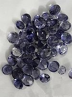 Photo showing a pile of approximately 46 small round faceted iolite gemstones of varying sizes and shades of bluish violet.