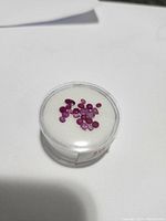 Top view of multiple genuine ruby gemstones in a round clear container on white cushion.