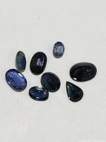 Eight loose sapphire gemstones of various shapes and shades of blue and dark tones laid on a white background.