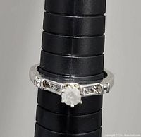 Close-up photo of a silver ring with a central round diamond and smaller side diamonds, displayed on a black ring sizing cone.