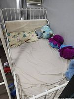 White metal twin bed frame with beige bedsheet, pillow with floral and bike pattern, and several stuffed toy animals on the bed.