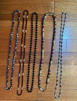 Top view of five various beaded necklaces arranged side by side on a wood floor.