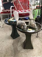 Two ceramic bird baths on pedestal bases with mesh bags of stones sitting inside. A box containing a solar water fountain device is on top of one bath.